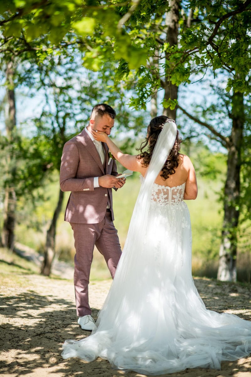 Bruid in witte kanten trouwjurk beïnvloedt liefje aan in mauve pak met briefje, intiem moment bij Garnwerd aan Zee onder bomen tijdens zonnige bruiloft.