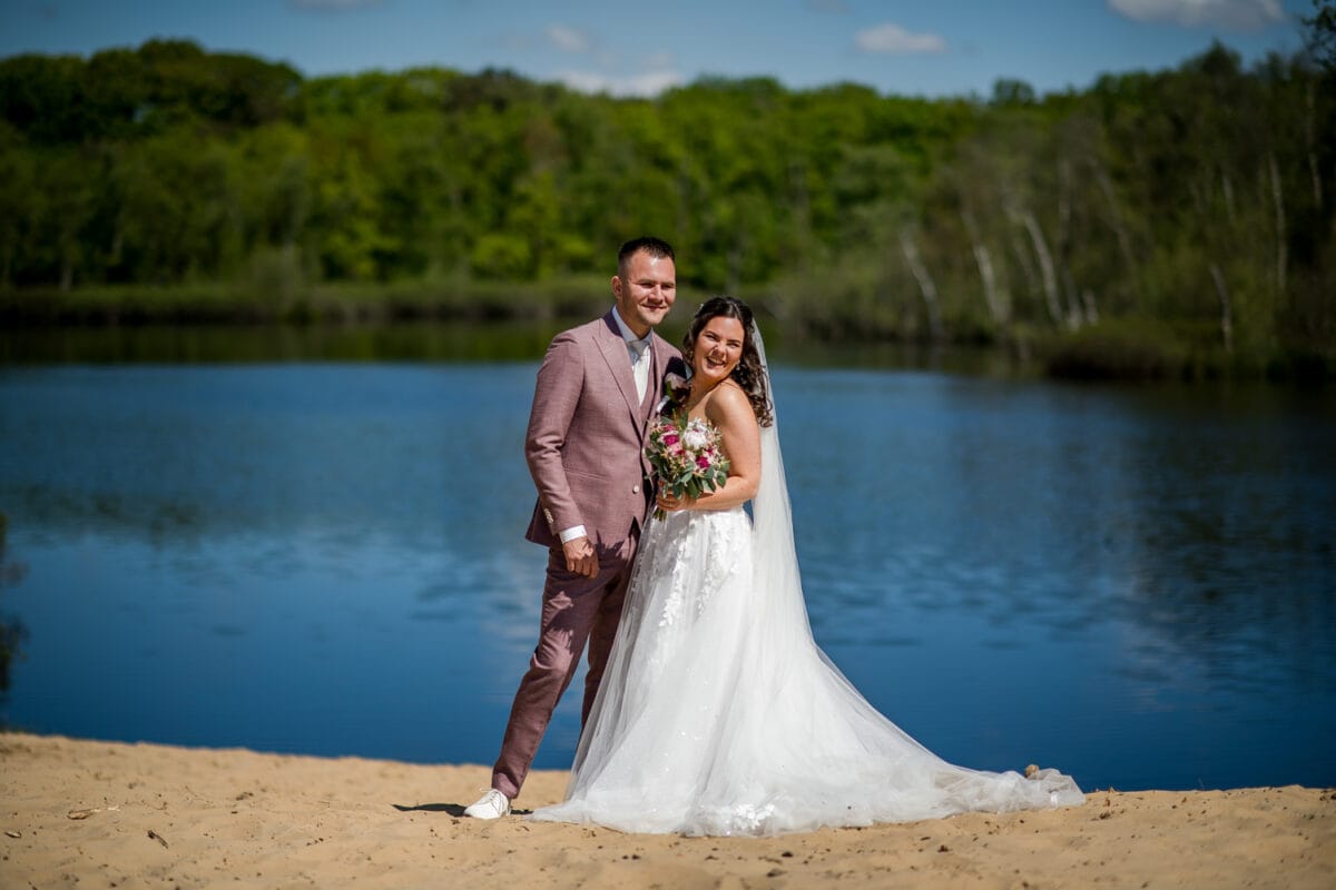 Bruid in witte jurk en sluier met boeket en bruidstaart in roze pak glimlachen op het strand van Garnwerd aan Zee, naast een helderblauw meer, groene bomen en zonlicht op de achtergrond.