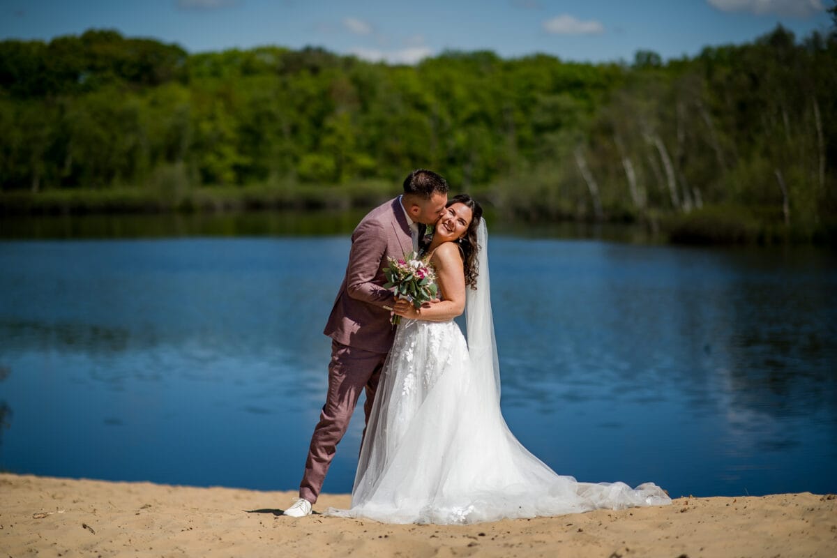 Bruidspaar bij Garnwerd aan Zee kust elkaar bij rustig meer, omringd door groene bomen, blauwe lucht en bruidsboeket. Romantische bruiloftsfoto in natuurlijk landschap.