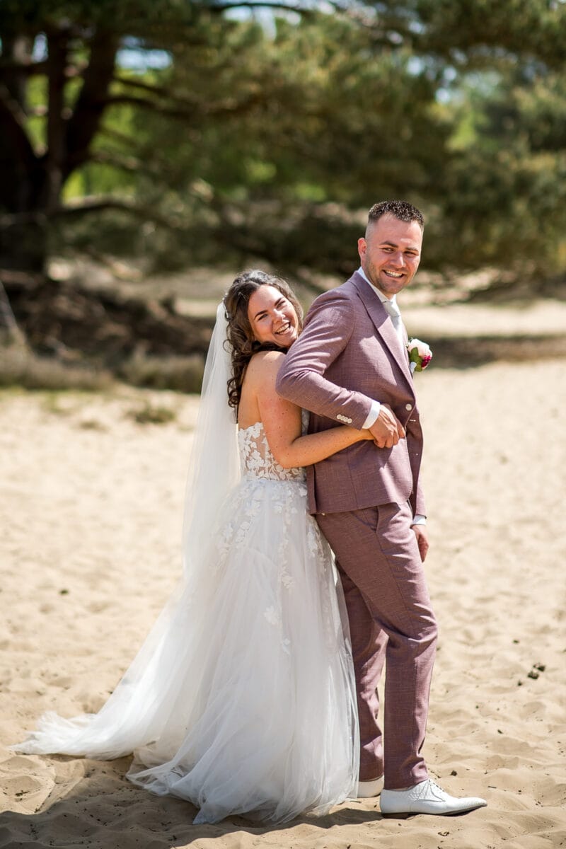 Bruid omhelst bruid op Garnwerd aan Zee bruiloft; zij in witte kanten trouwjurk, hij in lichtpaarse trouwpak en sneakers; zandgrond en groene bomen op zonnige dag.