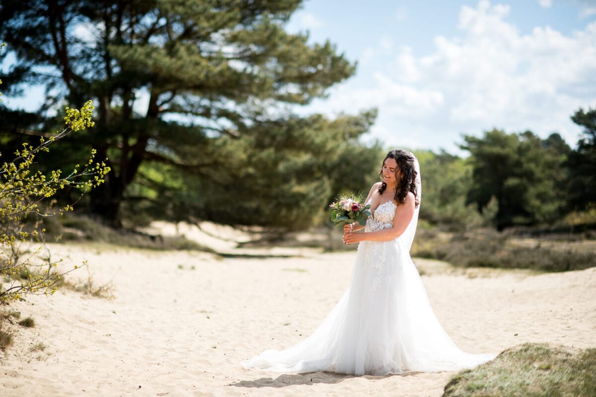 Bruid in witte trouwjurk met boeket op zandgrond buiten, verborgen door groene bomen en blauwe lucht met wolken, zonnige bruiloft sfeer, natuurlijke trouwlocatie.