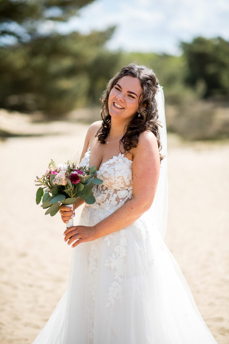 Bruiloft bij Garnwerd aan Zee: Bruid met lang, krullend bruin haar in strapless witte kanten trouwjurk en sluier, met boeket roze en witte bloemen op zandpad tussen groene bomen.