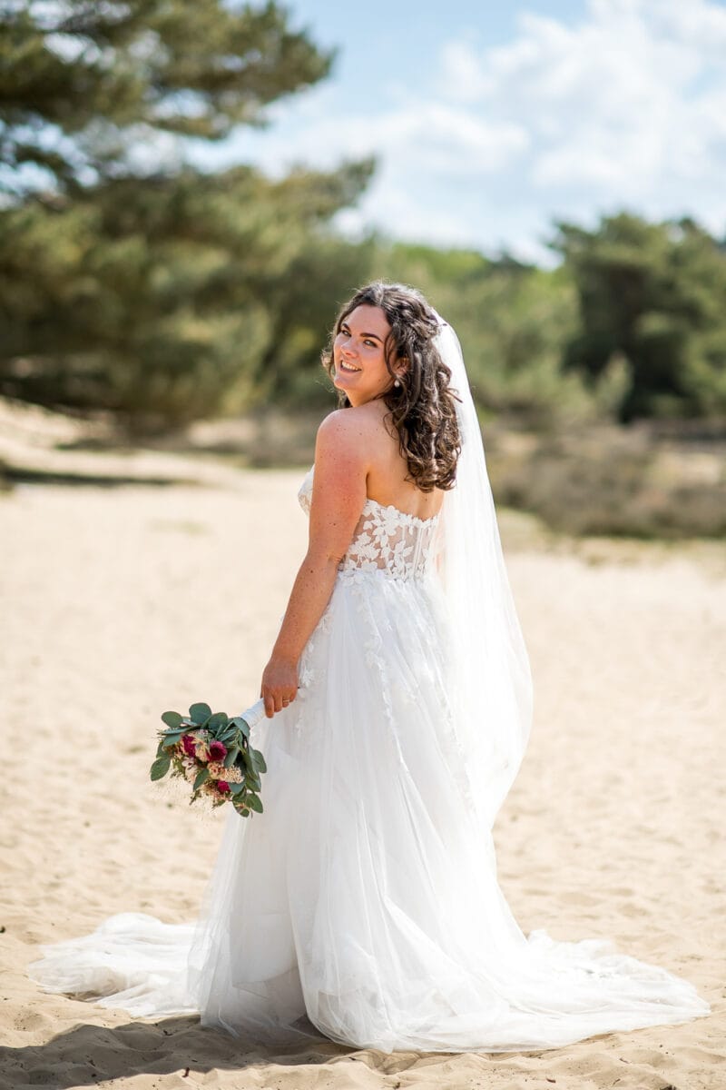 Bruiloft Garnwerd aan Zee: lachende bruid met lang bruin haar in strapless witte kant trouwjurk en sluier, boeket met rood en groen, zonlicht op buitenlocatie strand.