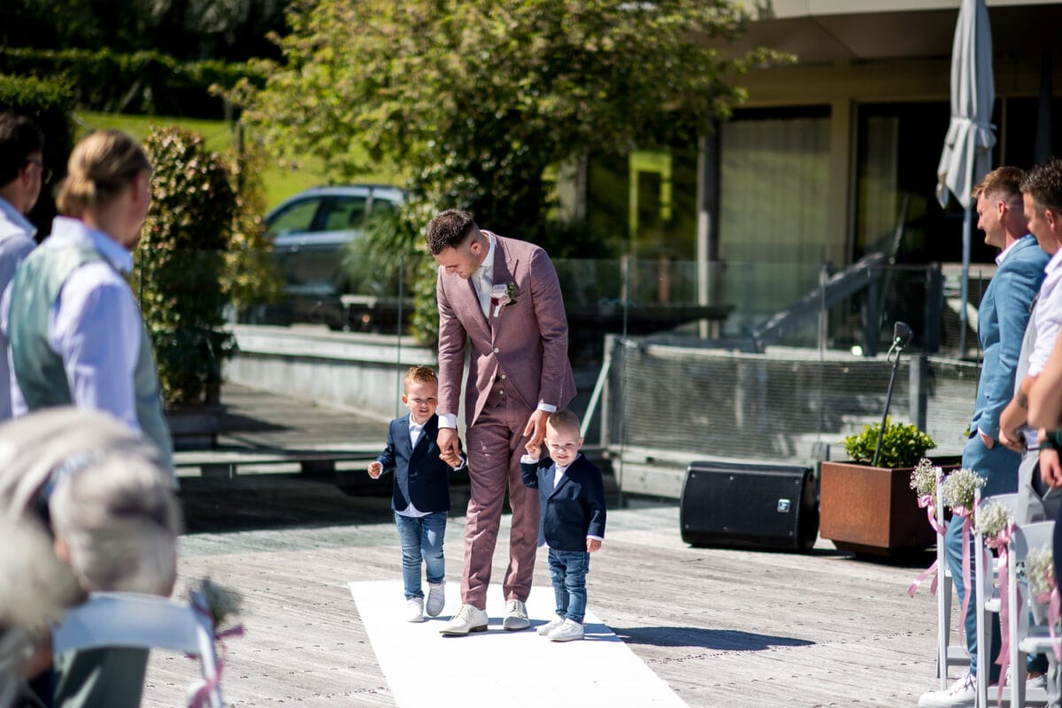 Bruiloft bij Garnwerd aan Zee: man in mauve pak loopt met twee jongens in blauwe jassen door buitentrouwlocatie tussen groen, gasten zitten naast het moderne gebouw.