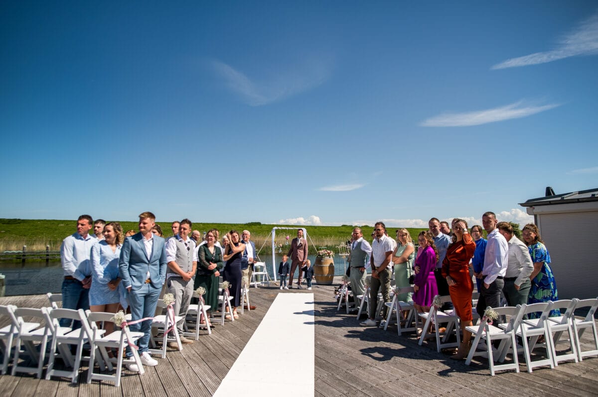 Bruiloft gasten op houten steiger bij Garnwerd aan Zee onder blauwe lucht, wachten bij witte loper op start huwelijksceremonie, sfeervolle buitenbruiloft.