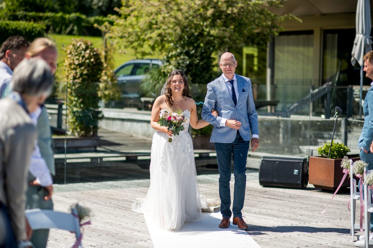 Bruid in witte trouwjurk loopt met begeleider over witte loper tijdens buitenbruiloft bij Garnwerd aan Zee, omringd door zonlicht en groen. Bruiloft fotografie sfeerimpressie.