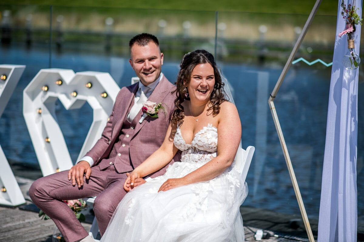 Bruid en bruid vieren hun bruiloft in Garnwerd aan Zee, beïnvloedend bij het water onder bloemenbogen en decoratieve letters, met zonlicht dat een feestelijke sfeer geeft.