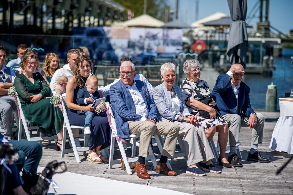 Bruiloft bij Garnwerd aan Zee met gasten in nette kleding op witte stoelen, stabiel weer, uitzicht op het water en een peuter samen met volwassene tijdens de huwelijksceremonie.