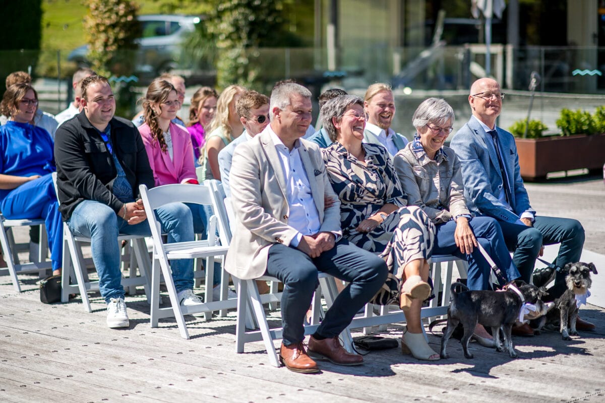 Bruiloft Garnwerd aan Zee: gasten op witte stoelen buiten, smart casual kleding, lachend met twee zwart-witte honden, zonnige en feestelijke sfeer met groen en gebouwen op de achtergrond.