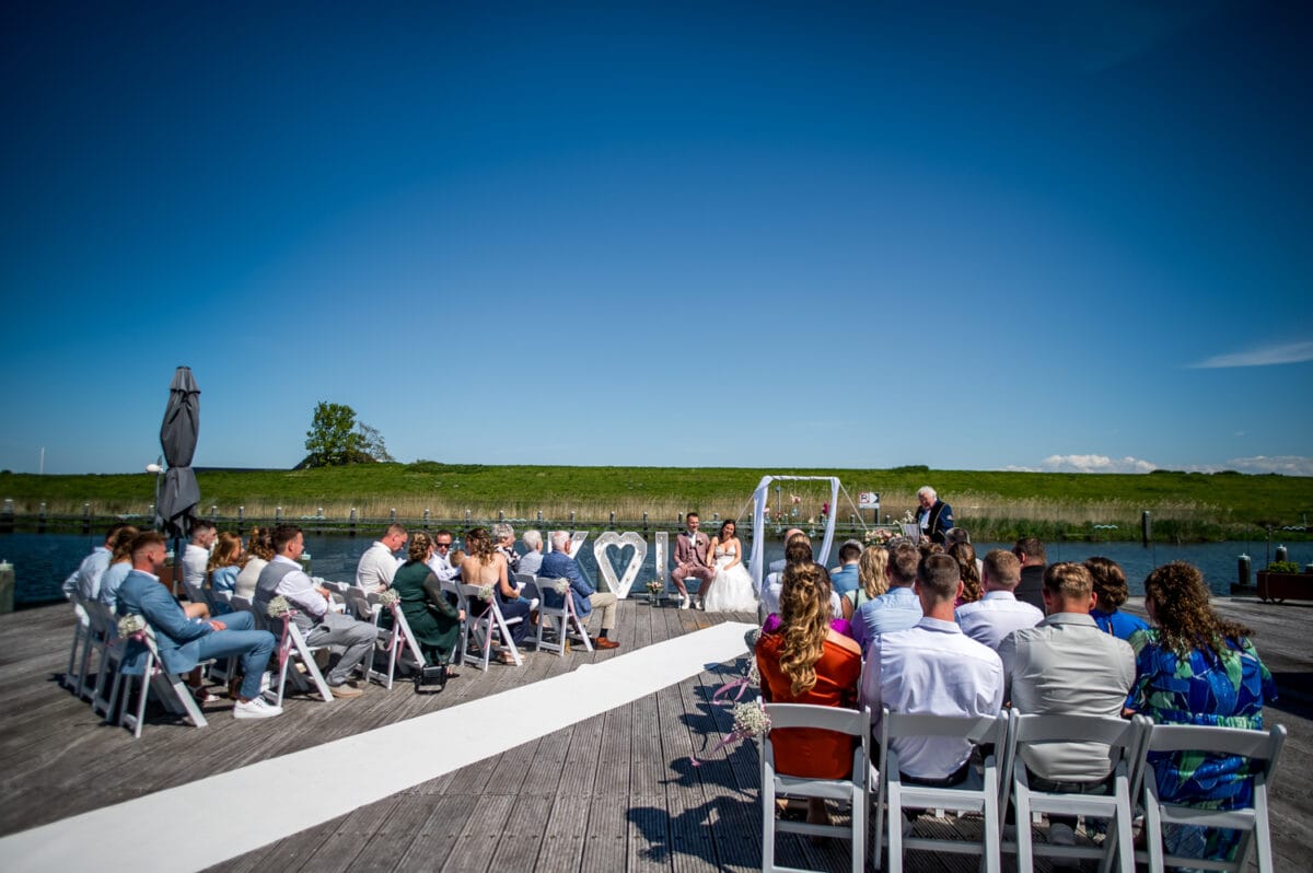 Buiten bruiloft bij Garnwerd aan Zee op een steiger, gasten op witte stoelen, moderne trouwprieel met hart decoratie, uitzicht op water en groene gras met bomen.