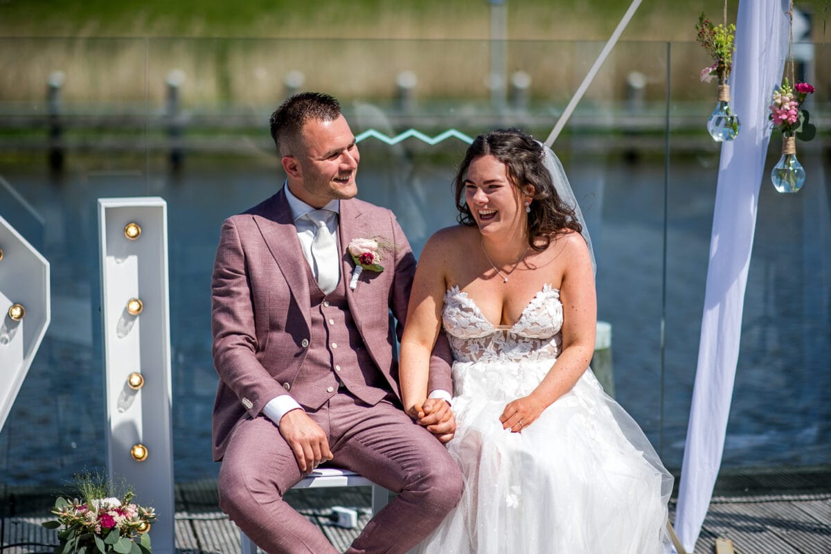 Bruidspaar zit hand in hand bij Garnwerd aan Zee, buiten aan het water. Bruiloft met zon, bloemen, glazen decoraties, witte trouwjurk met kant en mauve trouwpak.