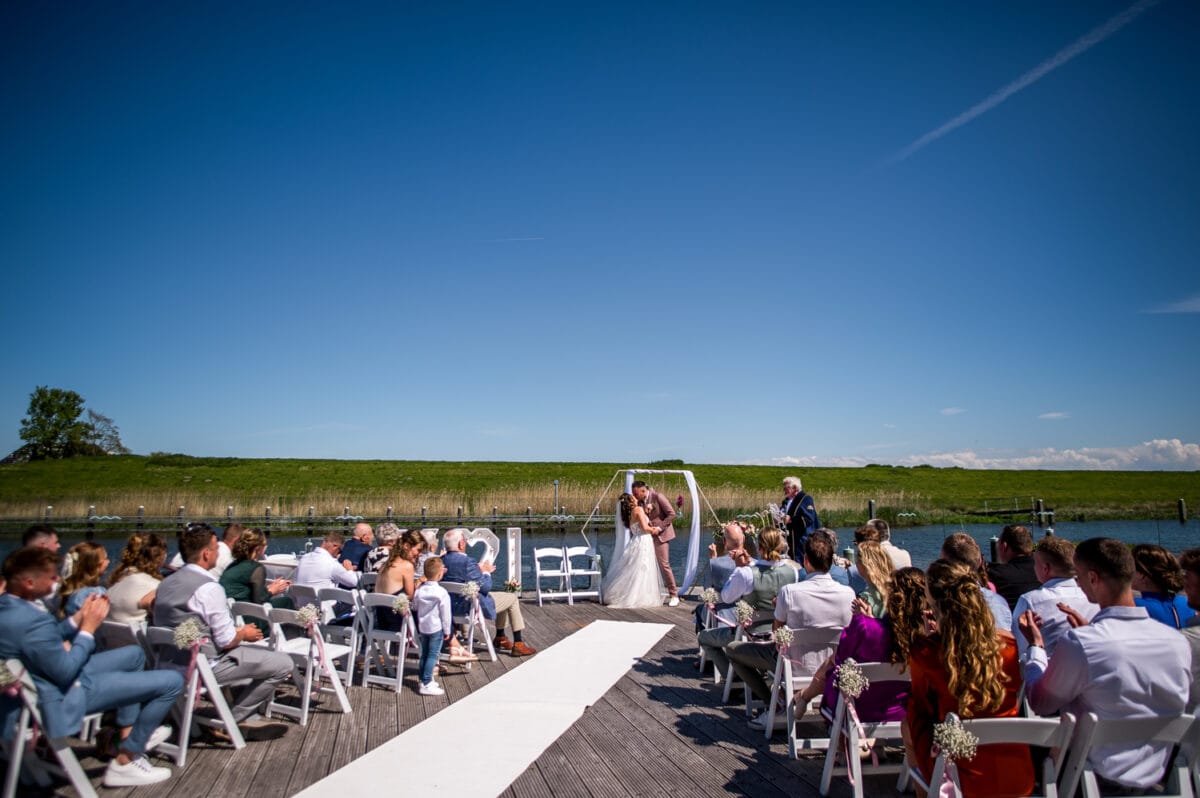 Buiten bruiloft ceremoniële boog aan het water bij Garnwerd aan Zee, bruidspaar en gasten op witte stoelen, blauwe lucht en groen gras, romantische trouwlocatie in Nederland.