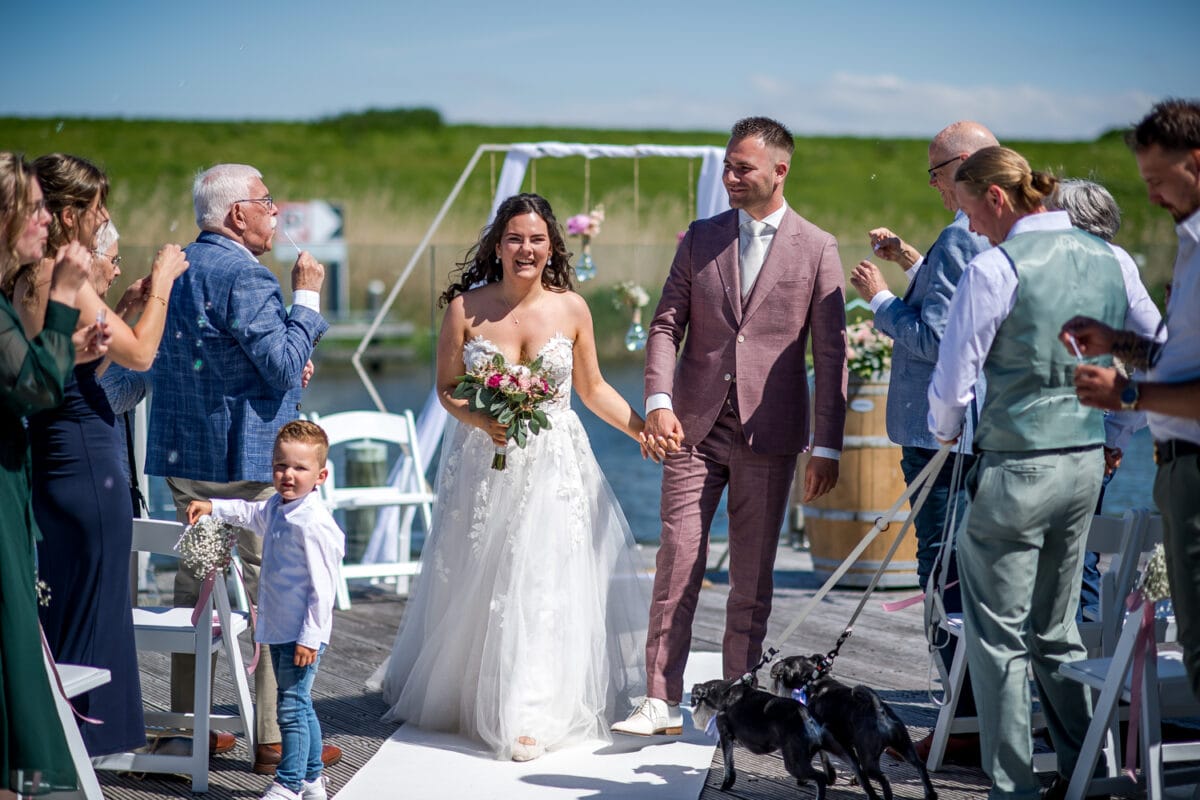 Bruid en bruidegom lopen hand in hand over het buitenpad tijdens hun bruiloft in Garnwerd aan Zee, met lachende gasten, twee kleine honden en krachtig weer tussen groene natuur.