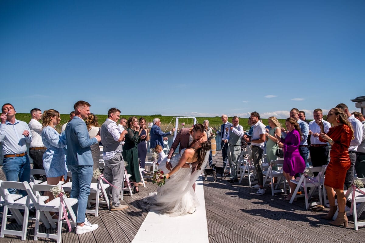 Bruid en bruid delen een romantische kus aan het einde van het buitenpad bij Bruiloft in Garnwerd aan Zee, omgeven door juichende gasten onder een succesvolle blauwe lucht.
