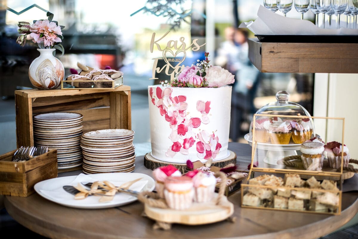 Rustieke desserttafel bij Garnwerd aan Zee bruiloft met witte taart met roze penseelstreek, cupcakes, gebak, glazen stolpen, houten kratten, servies en roze bloemen op ronde houten tafel.