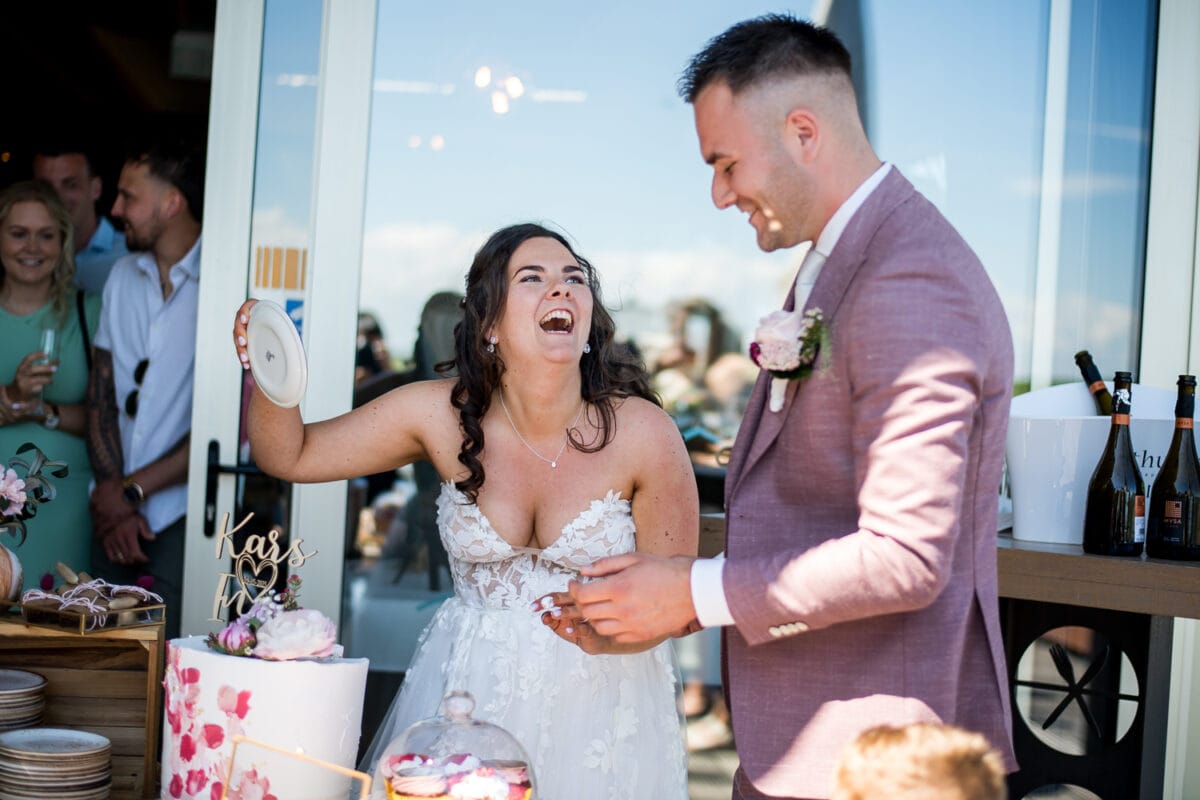 Bruid in witte kanten trouwjurk lacht naast pastelkleurige bruidstaart tijdens zonnige bruiloft bij Garnwerd aan Zee, trouwjurk in mauve pak, gasten vieren buitenfeest.