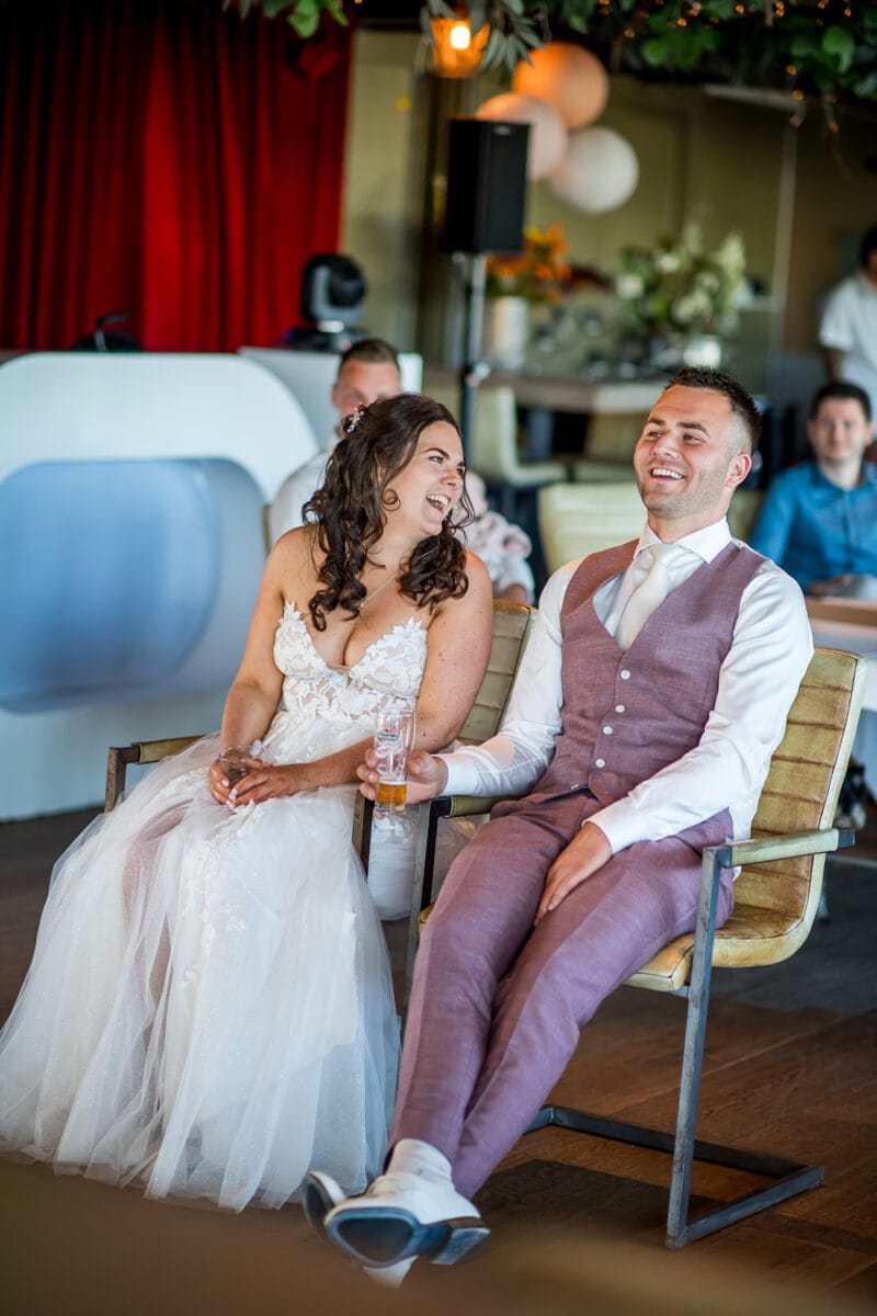 Bruiloft in Garnwerd aan Zee: bruid in witte kanten trouwjurk en bruid in mauve pak, hand in hand, rode gordijnen, ballonnen en feestelijke sfeer op trouwlocatie.