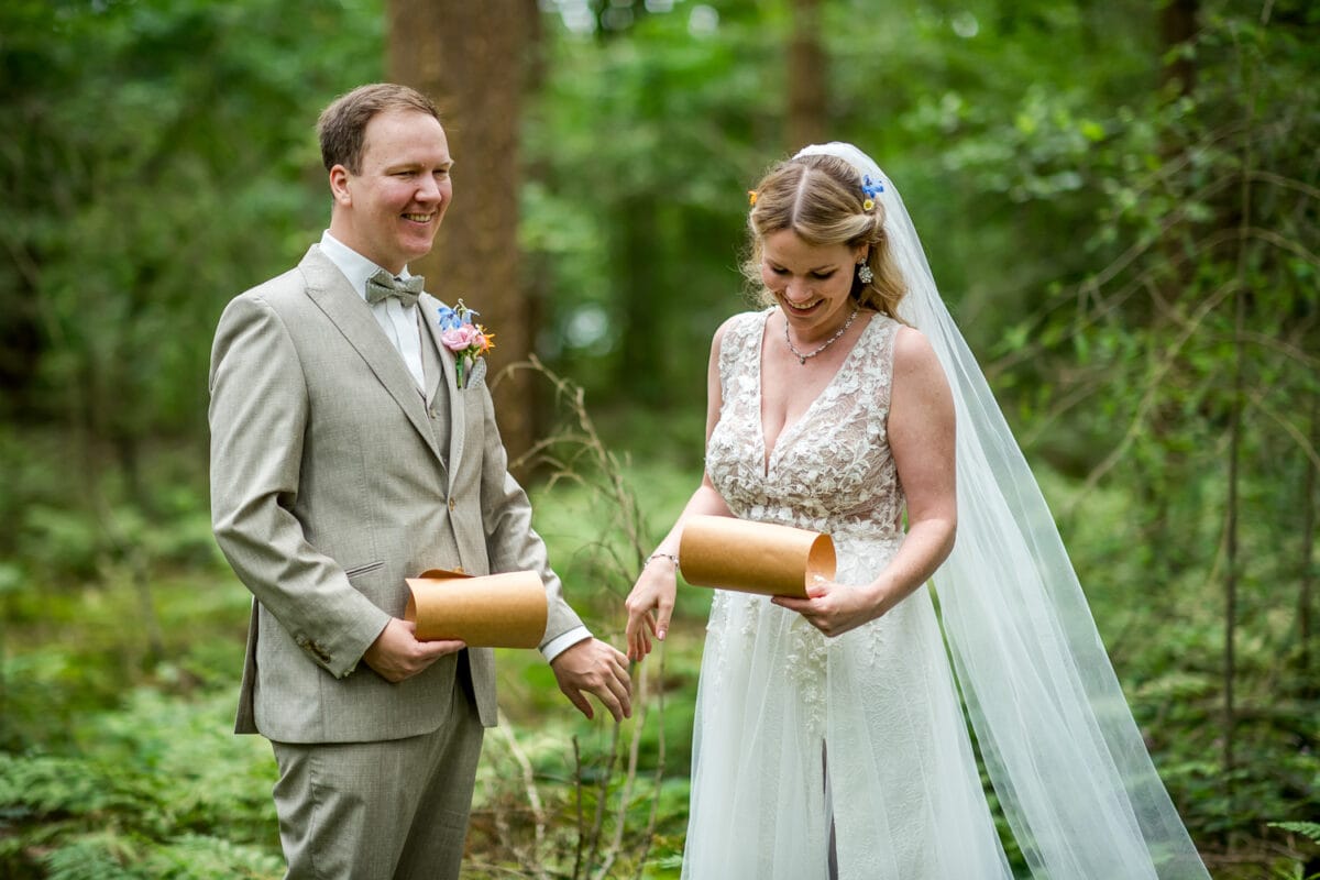 Stralend bruidspaar in groen bos, zij in witte kanten jurk met sluier, hij in licht beige pak, beiden lachend met gouden rollen.