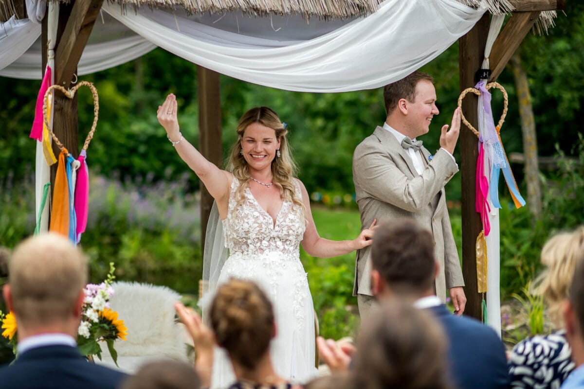 Bruiloftspaar onder versierde gazebo, zwaaiend naar gasten. Bruid in witte bloemenjurk, bruidegom in beige pak, kleurrijke decoratie.
