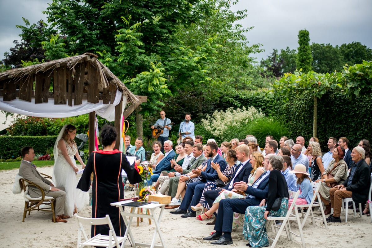 Rustieke buitenbruiloft bij de Kruimel, stel onder een overkapping, muzikanten spelen gitaar, gasten in feestelijke kleding.