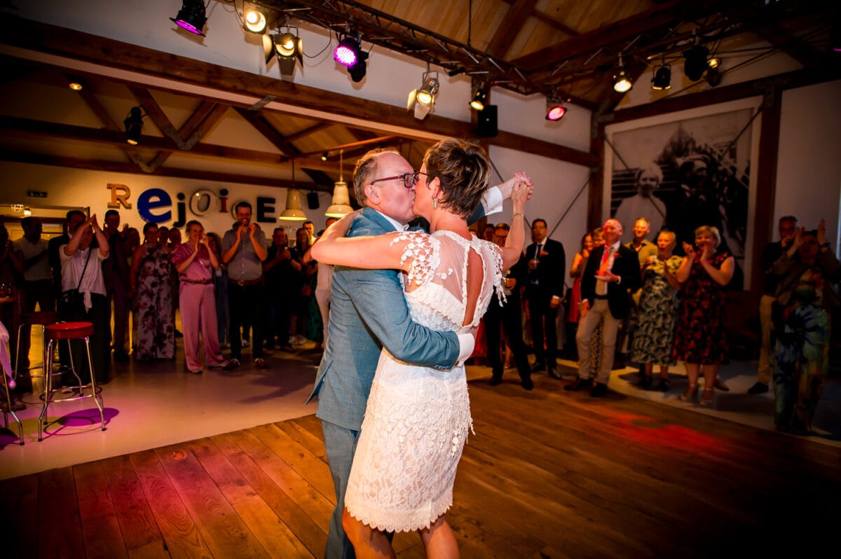 Bruidspaar deelt kus tijdens bruiloft in Rietland, dansend onder warme verlichting, gasten applaudisseren in feestzaal.