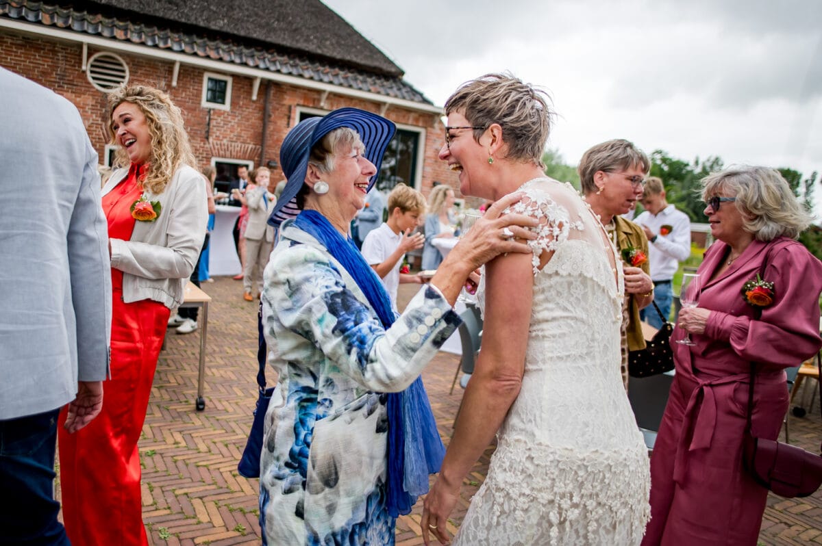 Twee vrouwen lachen en omhelzen op een bruiloft bij Rietland, gasten in kleurrijke kleding op een bakstenen terras bij een landelijke locatie.