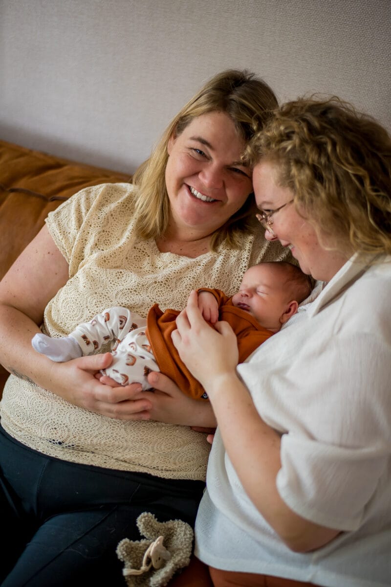 Twee vrouwen op bank glimlachend, houden slapende baby in oranje kleding vast, tonen genegenheid en vreugde in een gezellige woonkamer.