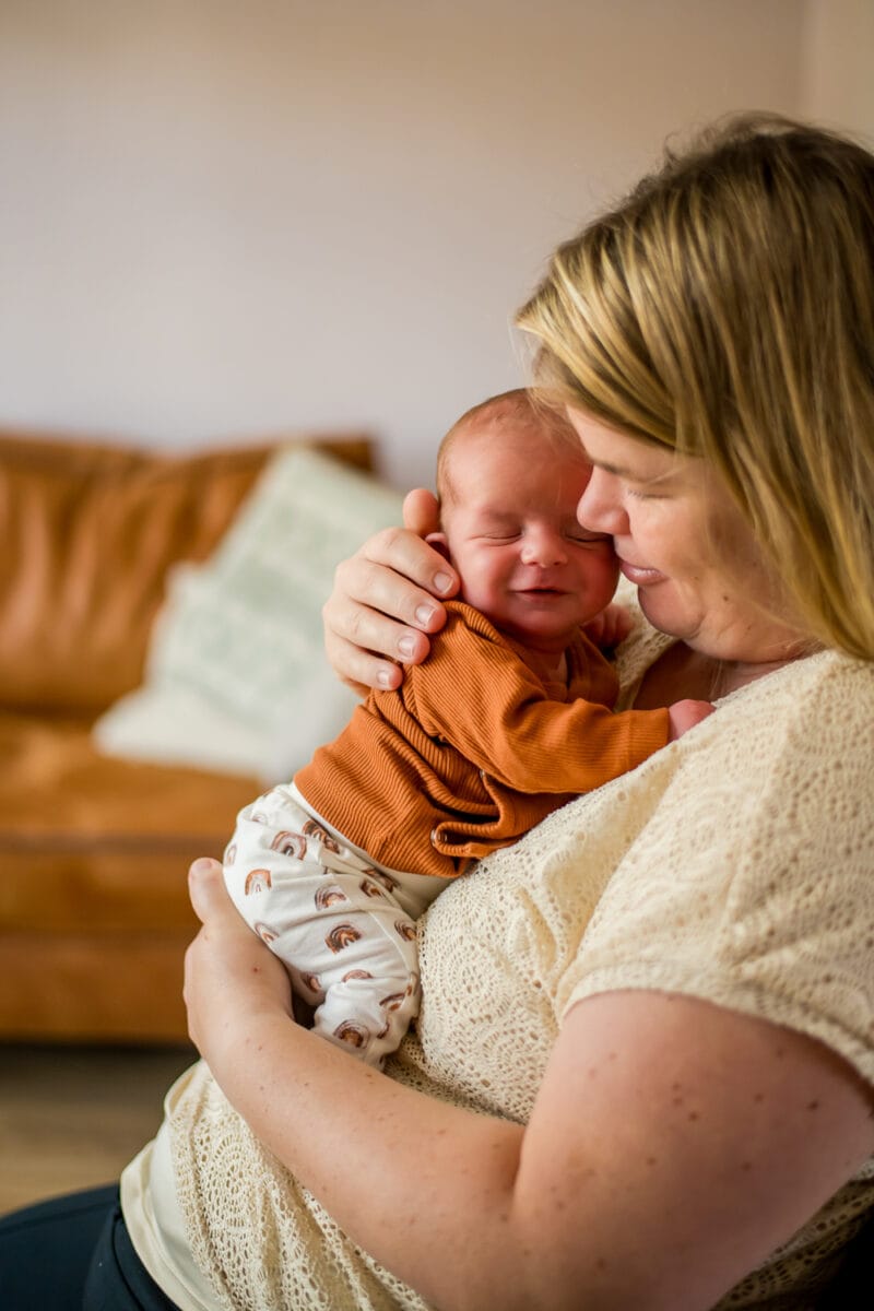 Blonde vrouw houdt pasgeboren baby met regenboogbroekje vast in knusse woonkamer met bruine bank en sierkussen.