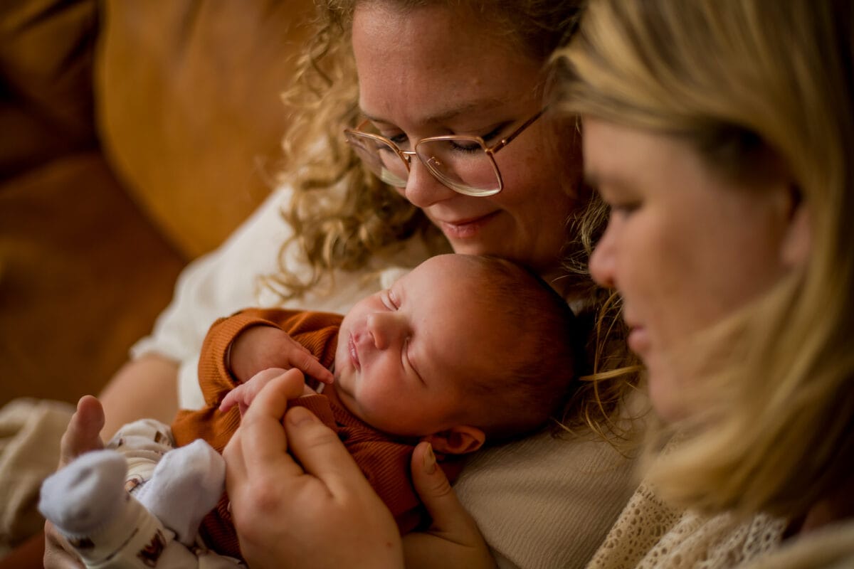 Twee vrouwen knuffelen een slapende pasgeborene in roestkleurige kleding en delen een teder, liefdevol familiemoment in warm, intiem licht.