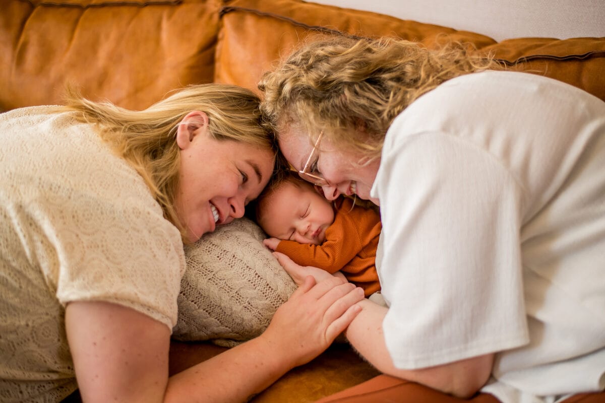 Twee volwassenen met blond haar buigen liefdevol over slapende baby in oranje op bruine bank, gezichten dichtbij, warme sfeer.