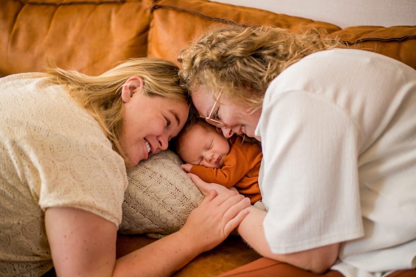 Twee volwassenen met blond haar buigen liefdevol over slapende baby in oranje op bruine bank, gezichten dichtbij, warme sfeer.