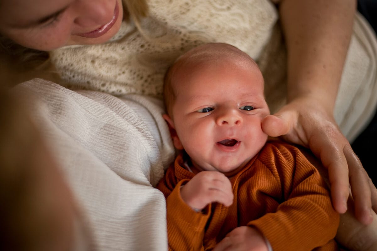 Vrolijke baby in roestkleurige outfit ligt in armen van volwassene in crème trui, intiem moment van liefdevolle band.