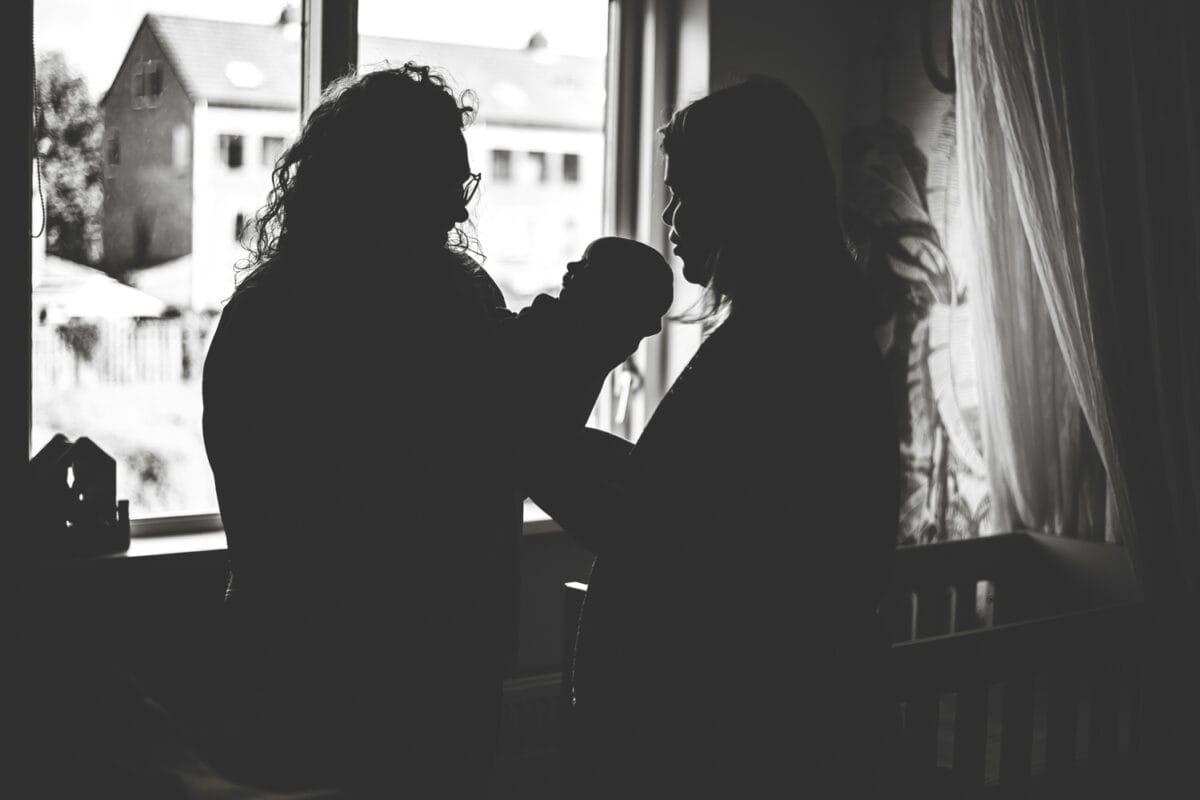 Twee silhouetten van volwassenen bij helder raam in donkere kamer, object wordt doorgegeven, vervaagde huizen en licht buiten.