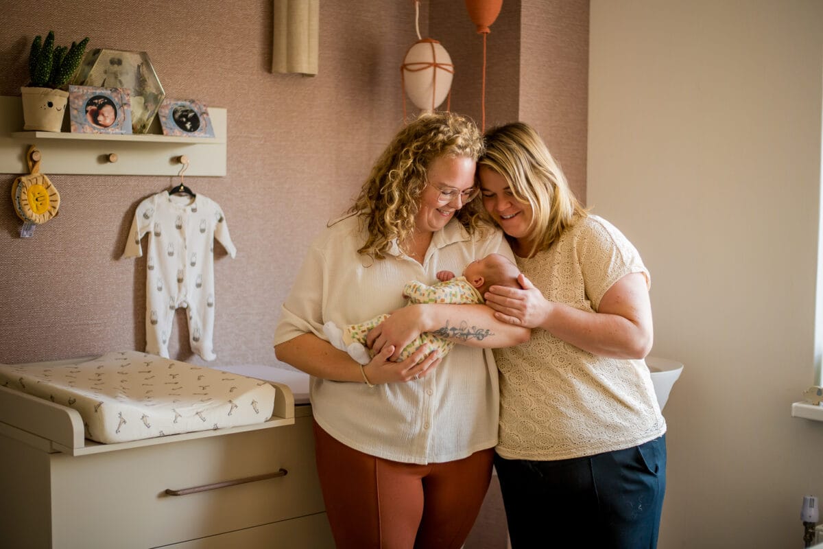 Stel met pasgeboren baby in knusse babykamer met warme verlichting, babykleding aan de muur en pasteltinten.