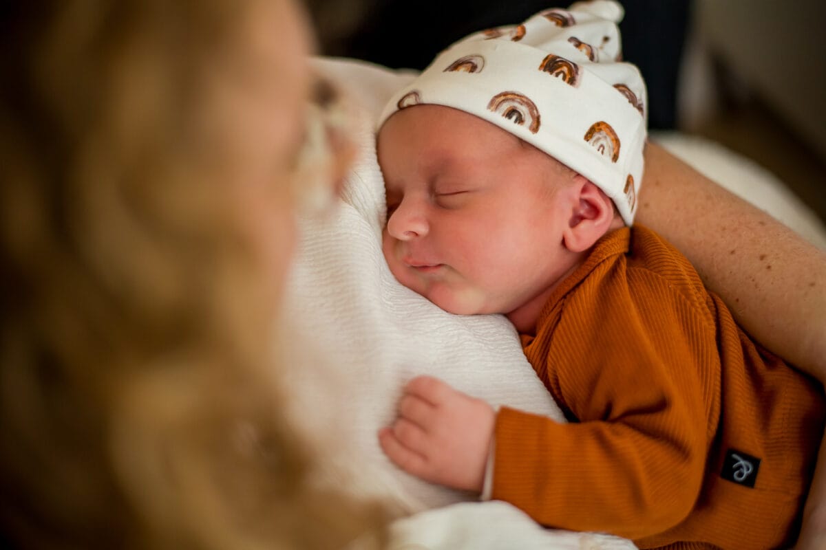 Pasgeboren baby in witte regenbooghoed en bruine outfit slaapt op de borst van een volwassene, gezellige omgeving, zachte omhelzing.