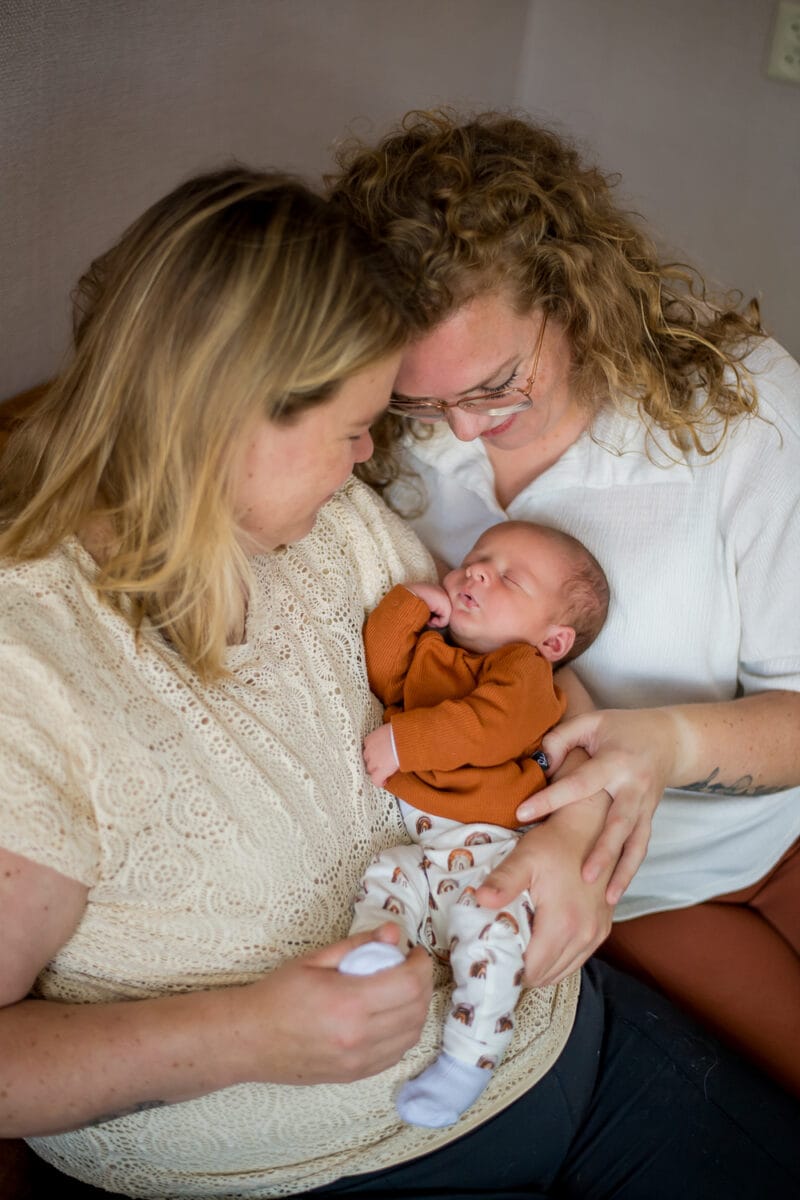 Twee vrouwen met licht haar glimlachen, houden een pasgeborene vast in een roestkleurig topje en een broek met patroon, en tonen liefde tijdens een intiem familiemoment.