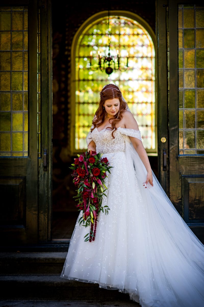 Bruid met lang rood haar in witte kanten off-shoulder jurk, parel hoofdband, houdt rood boeket vast voor sierlijke glas-in-lood deuren.