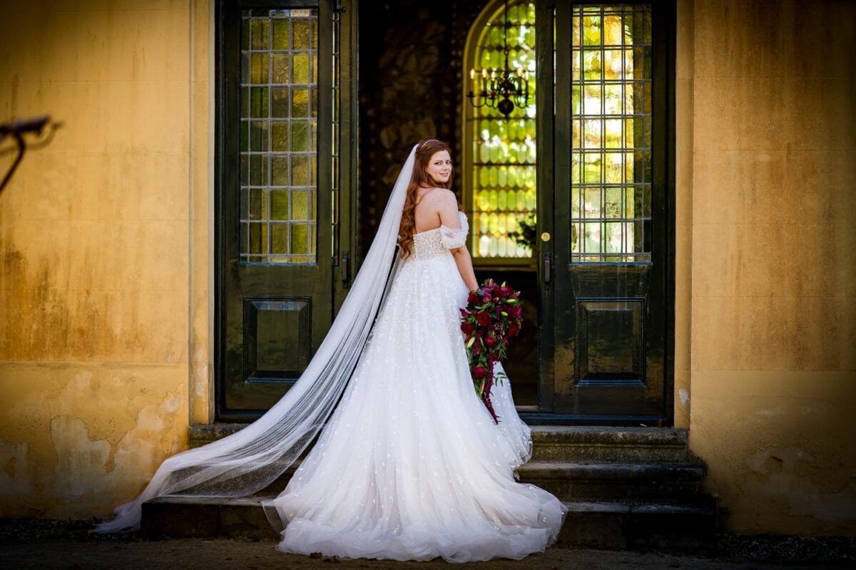 Bruid met lang rood haar in off-shoulder bruidsjurk en sluier houdt rood boeket vast op stenen trap voor groene deuren, bruiloftssetting.
