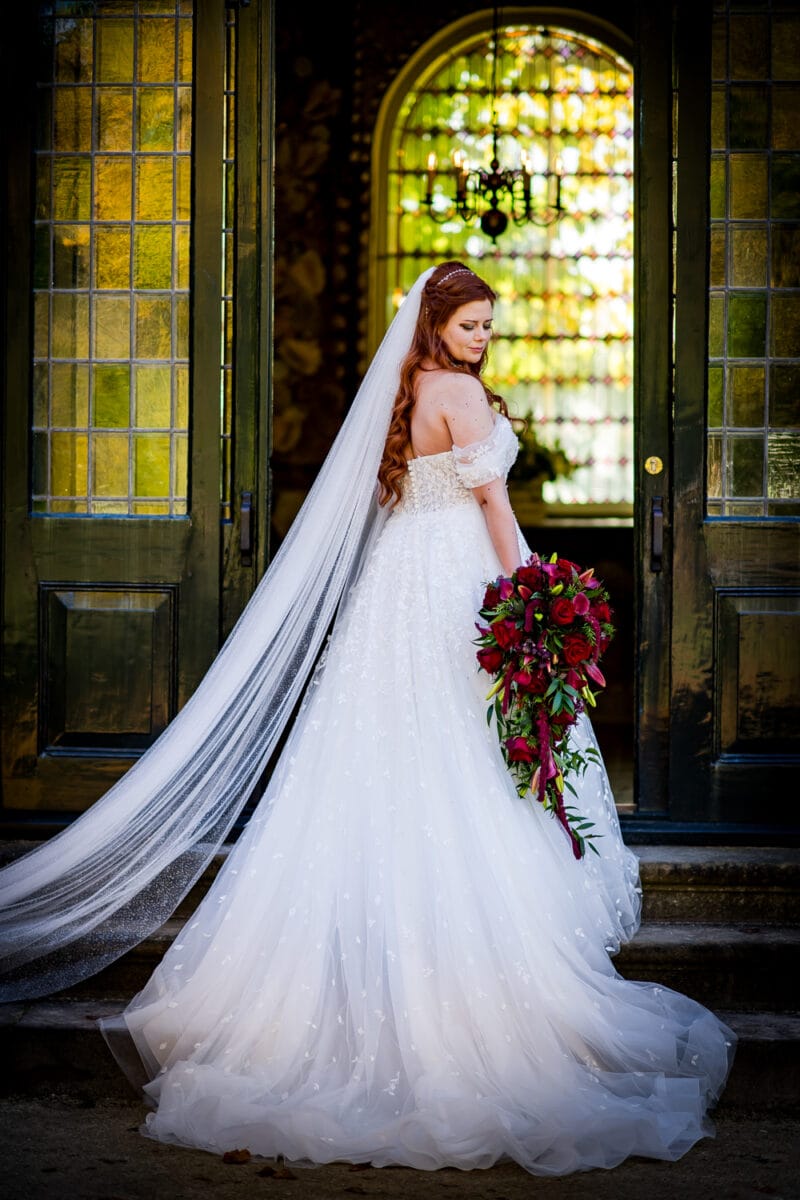 Bruid in witte off-shoulder trouwjurk met lange sluier en rood rozenboeket op stenen trap, zonlicht door boograam.