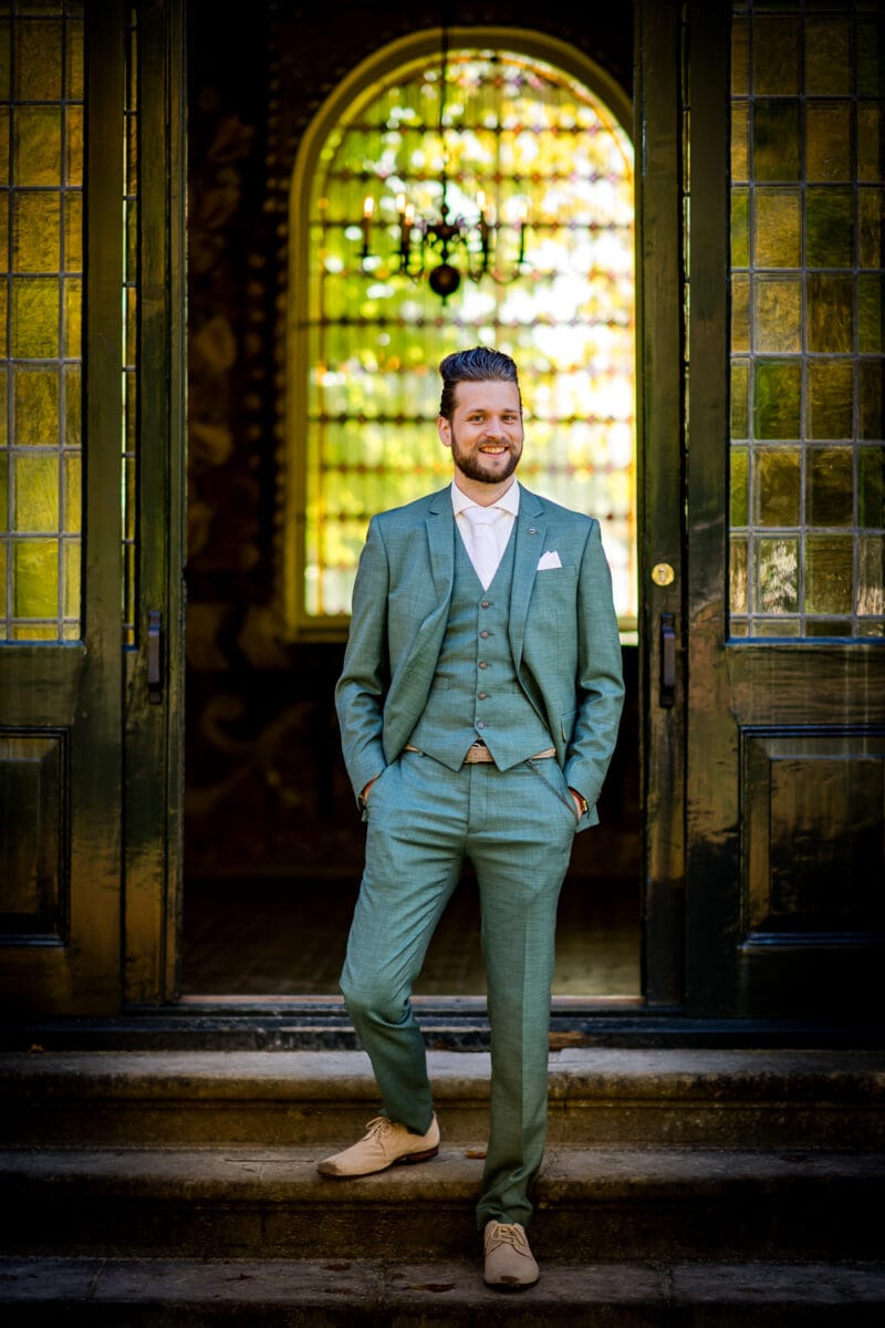 Man met baard in licht groenblauw pak op stenen trap, geflankeerd door open deuren, met oplichtend gebogen raam in glas-in-lood op de achtergrond.