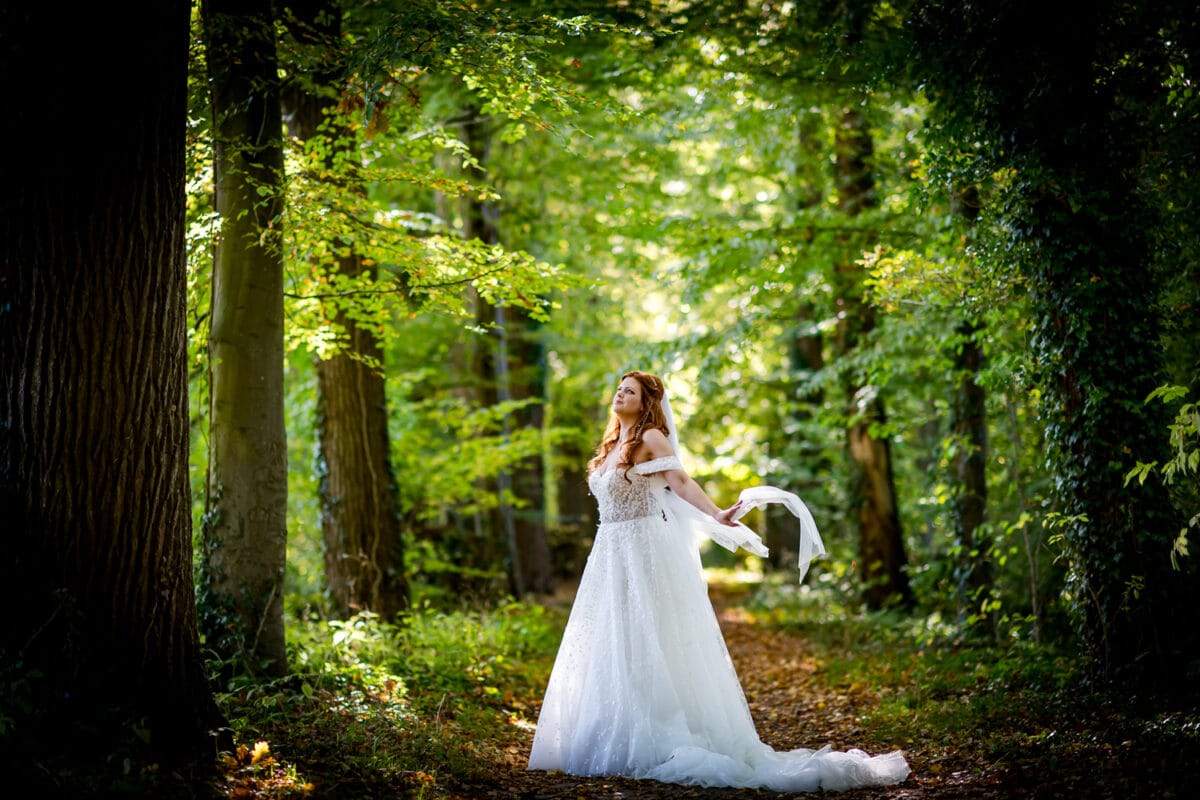 Bruid in witte off-shoulder trouwjurk op bospad, zonlicht door bomen, met doorzichtige sjaal, dromerige serene sfeer.