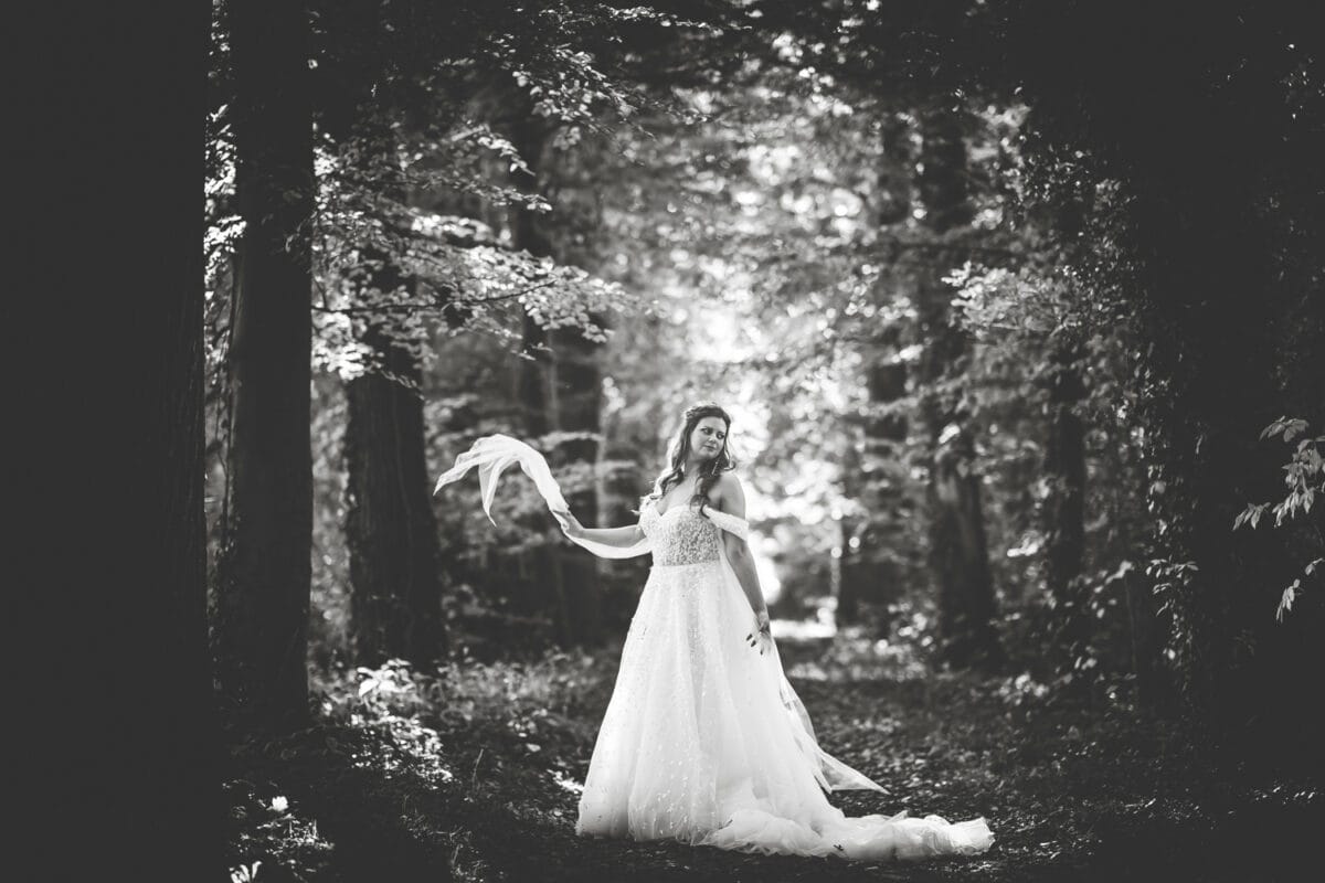 Bruid in vloeiende witte jurk in zonovergoten bos, sluier vasthoudend, zacht glimlachend. Dromerige, romantische zwart-wit trouwfoto.