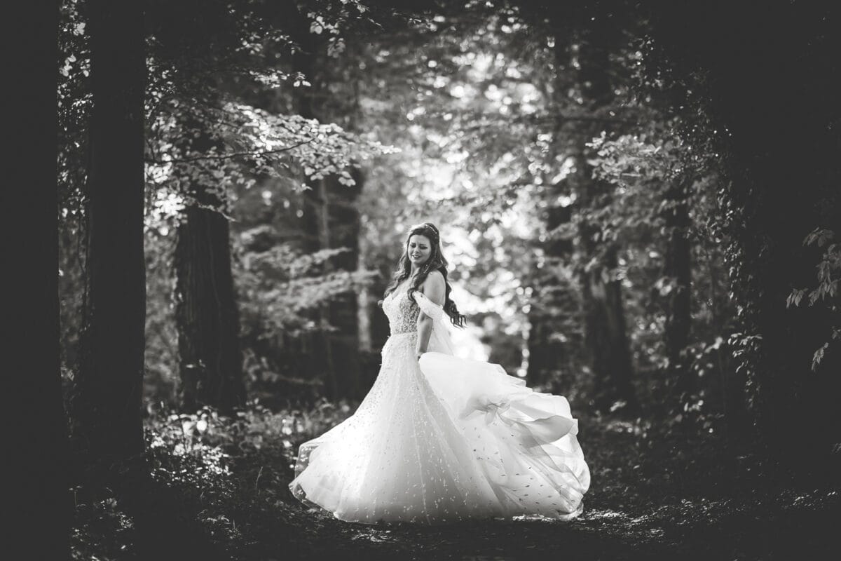 Twirling bruid in versierde trouwjurk in zonovergoten bos, hoge bomen, romantische zwart-wit bruidsfoto, dromerige sfeer.