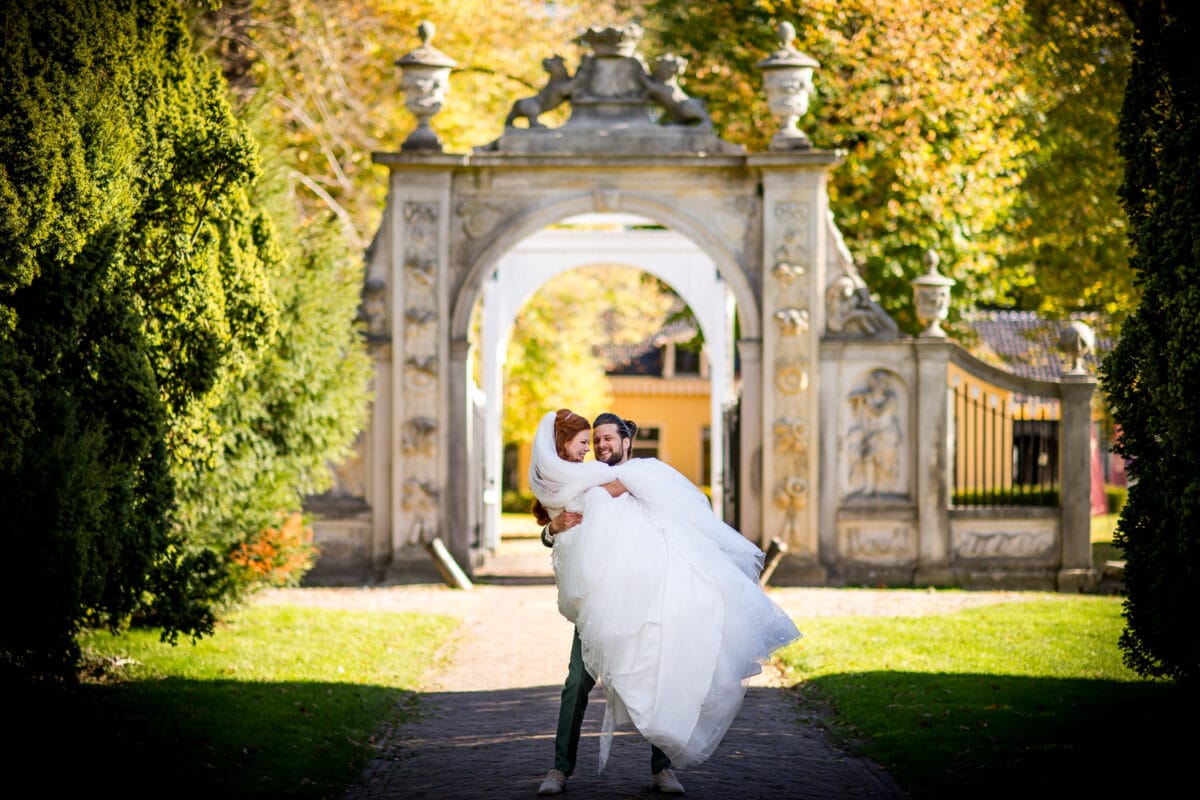 Bruidegom in grijs pak draagt bruid in witte jurk op zonovergoten pad, omringd door groen, boog en gele trouwlocatie.