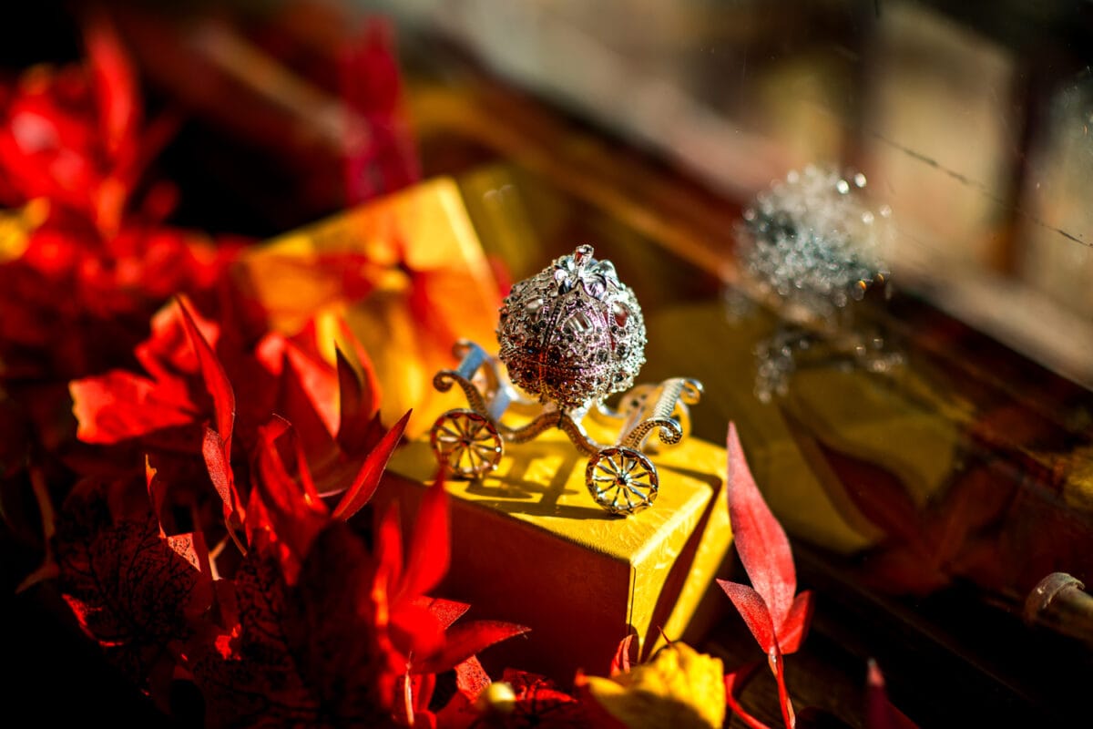 Zilveren filigraan pompoenkoets op gele doos bij raam, omringd door rode herfstbladeren, zonlicht reflecteert, warme sfeer.
