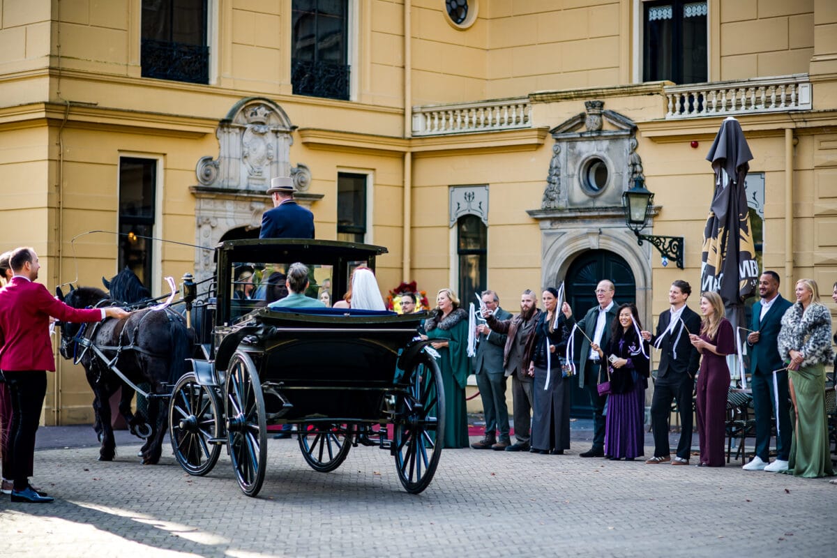Zwarte paardenkoets verlaat historisch geel gebouw, goed geklede gasten buiten vieren bruiloft of feestelijke gebeurtenis.