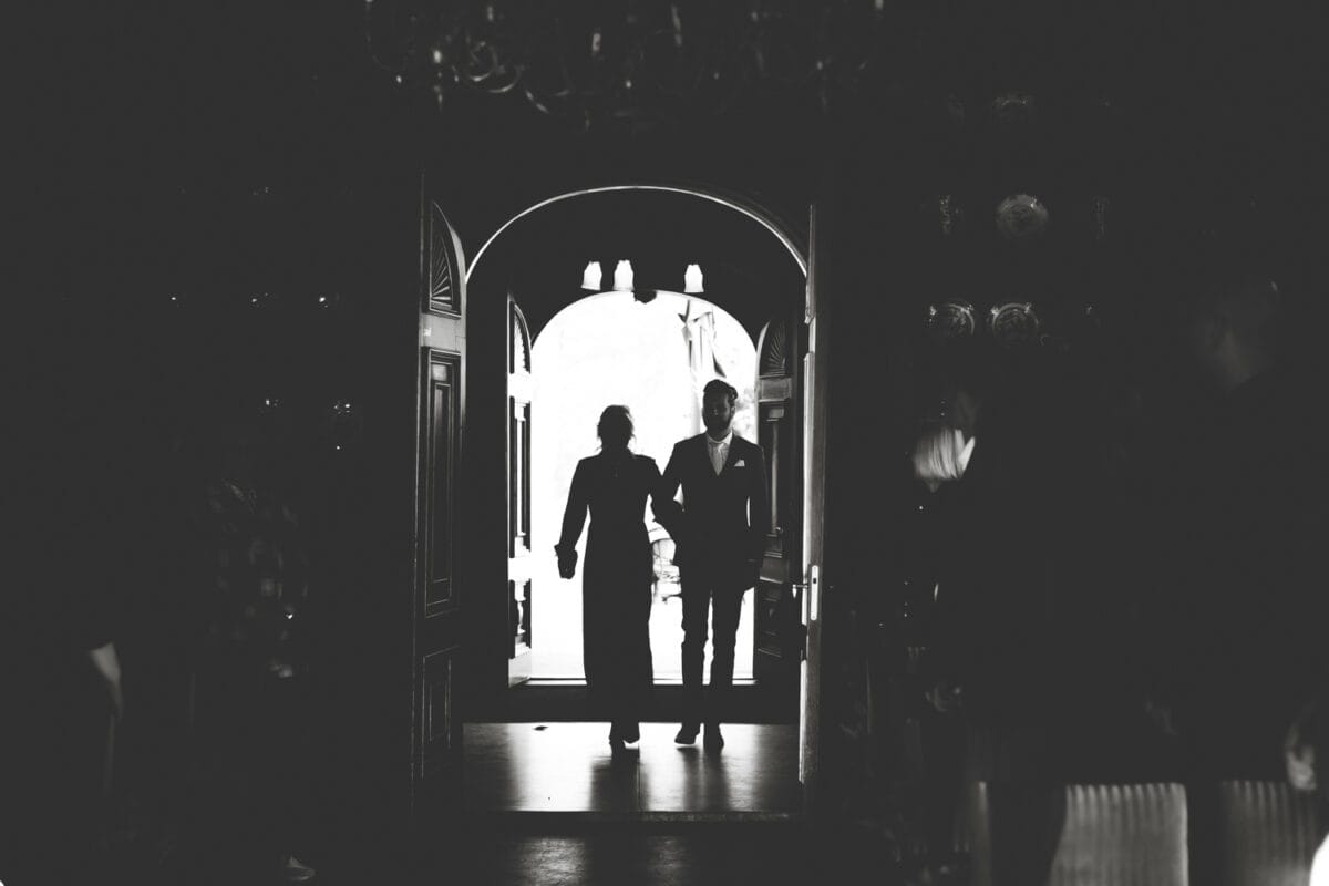 Zwart-wit foto van paar in nette kleding, lopend door grote open deur naar donkere ruimte, silhouetten door fel daglicht.