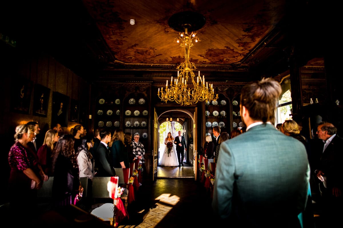 Bruid in witte jurk en sluier loopt met vader door het gangpad in grote, met kroonluchters verlichte trouwlocatie terwijl gasten naar de stoet kijken.