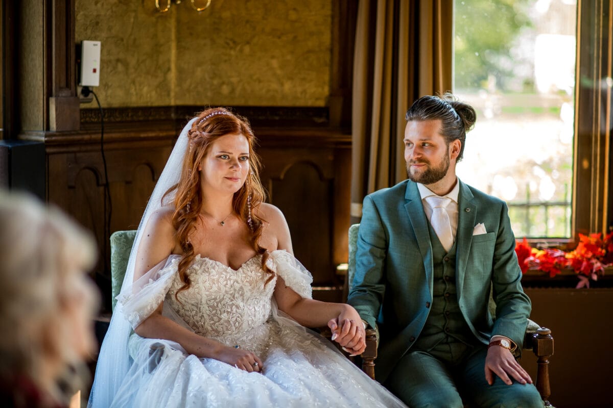 Bruiloftspaar binnen, bruid met witte off-shoulder jurk en sluier, bruidegom in groen pak, herfstdecoratie, zonlicht.