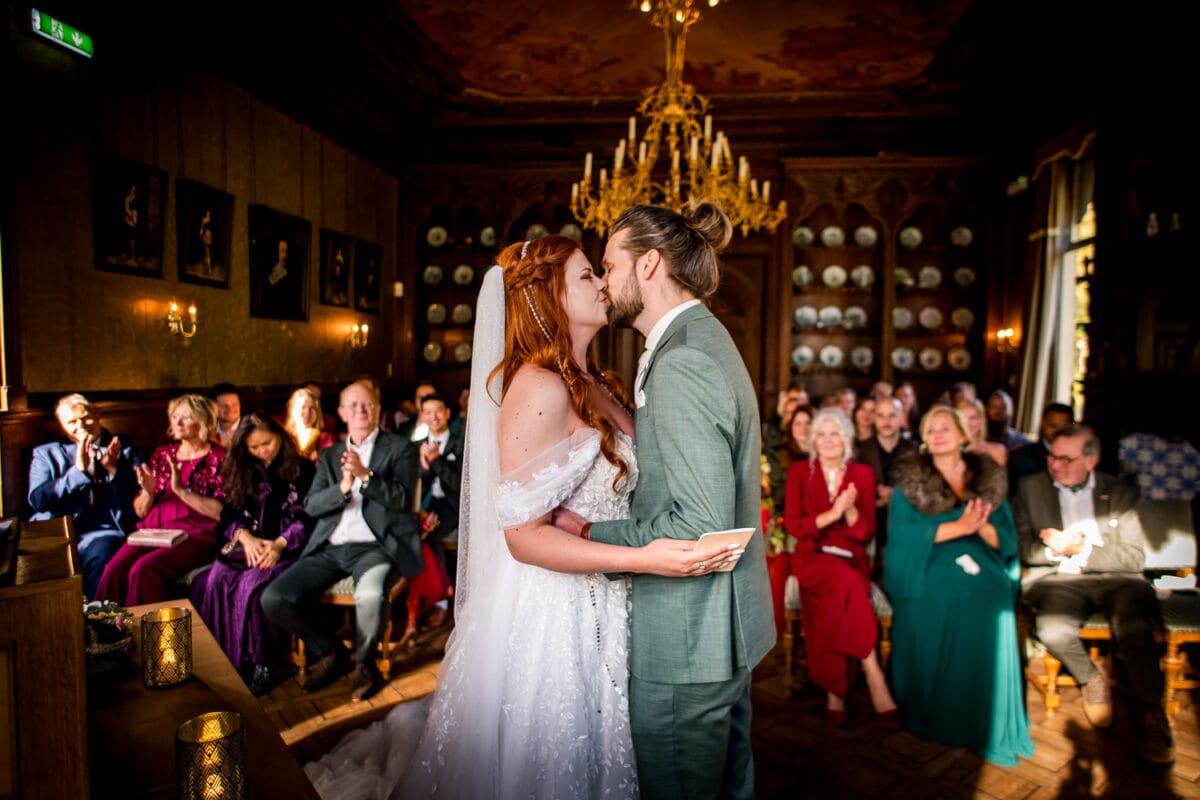 Huwelijksceremonie: bruid in witte jurk en bruidegom in lichtgroen pak kussen elkaar, sierlijke kamer, gasten applaudisseren, kroonluchter zichtbaar.